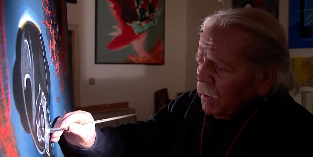 Eine ältere Person mit grauem Haar und Schnurrbart malt in einem schwach beleuchteten Raum auf einer Leinwand. In der rechten Hand hält sie einen Pinsel. An der Wand hinter ihr hängen farbenfrohe Kunstwerke.