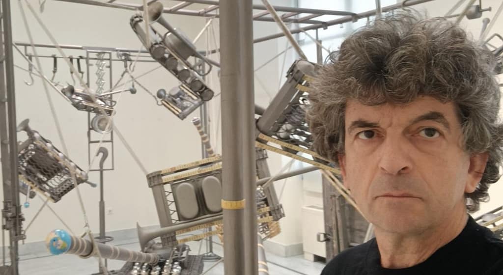 Ein Mann mit lockigem Haar steht in einem Galerieraum vor einer großen, komplexen Metallkunstinstallation mit verschiedenen mechanischen und industriellen Objekten, die an Stangen und Drähten aufgehängt sind.