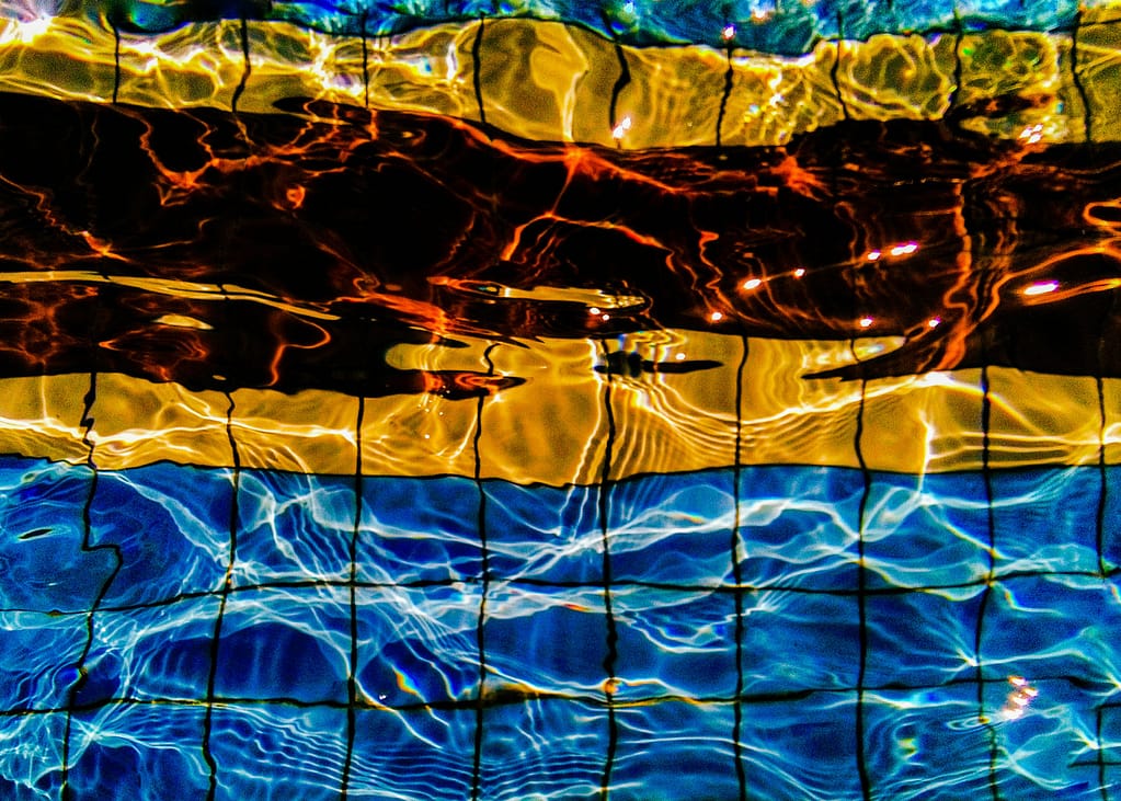 Farbenfrohe, abstrakte Ansicht von Poolwasser mit einem Gittermuster; Sonnenlicht erzeugt leuchtende blaue, gelbe und orangefarbene Reflexionen und wellenförmige Verzerrungen auf dem Poolboden.