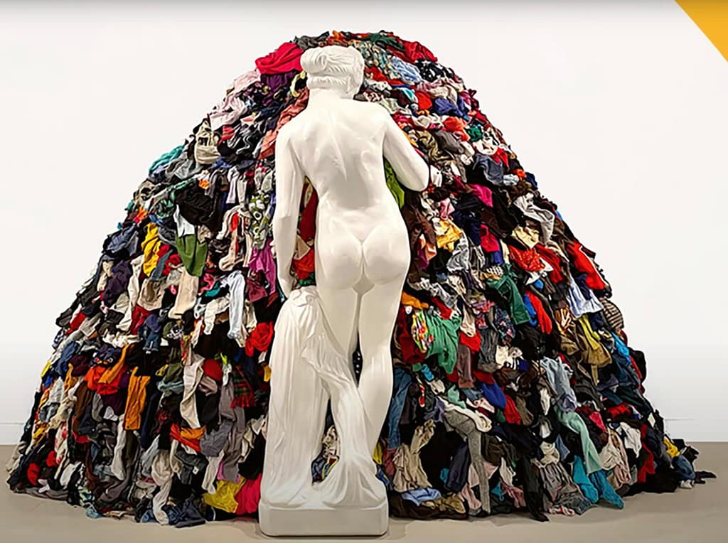 In einer Kunstgalerie mit schlichter weißer Wand steht die weiße Marmorstatue einer nackten Frau vor einem großen Haufen bunter, aufgestapelter Kleidung.