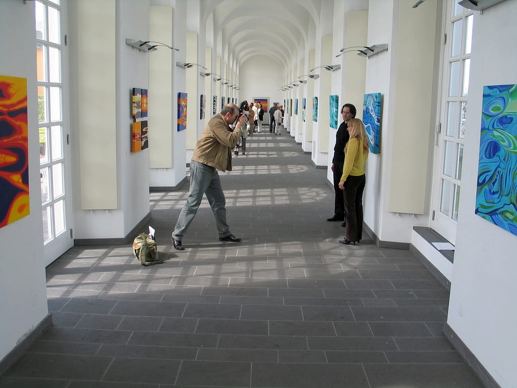 Ein Mann macht ein Foto von einer Frau in einem hellen, gewölbten Flur, der mit farbenfrohen abstrakten Gemälden gesäumt ist; im Hintergrund gehen oder stehen mehrere Personen.