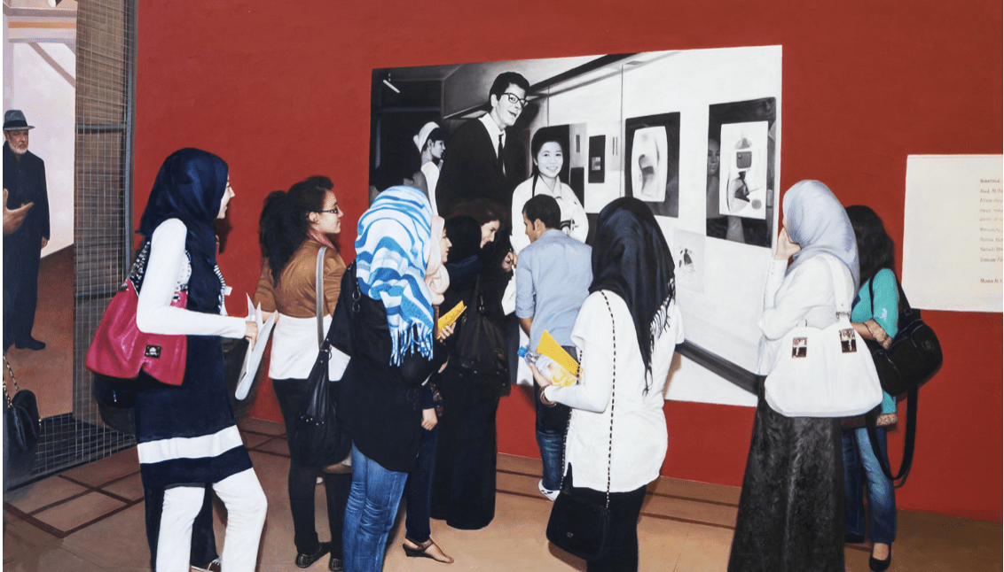 Eine Gruppe versammelt sich vor einer Wand, an der große Fotografien einer Ausstellung mit dem Titel „Positionen zur Malerei“ hängen. Einige tragen Hijabs, andere halten Broschüren in den Händen. Die rote Wand und der rote Boden bilden einen Kontrast zu einer schwarz gekleideten Person, die daneben steht und dieser künstlerischen Szene Tiefe verleiht.