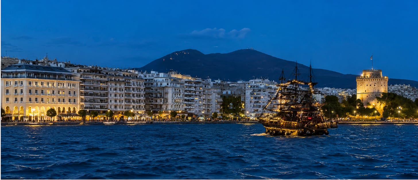 Die Uferpromenade der Stadt in der Abenddämmerung, mit einem hell erleuchteten historischen Schiff auf dem Wasser, beleuchteten Gebäuden entlang des Ufers und einem runden Steinturm mit einer griechischen Flagge im Hintergrund.