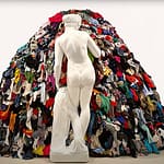 In einer Kunstgalerie mit schlichter weißer Wand steht die weiße Marmorstatue einer nackten Frau vor einem großen Haufen bunter, aufgestapelter Kleidung.