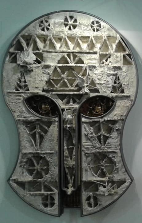 Eine metallische, abstrakte Maskenskulptur mit geometrischen Mustern und Ausschnitten, die zwei markante Augenformen und eine verwitterte, strukturierte Oberfläche in Grau- und Weißtönen aufweist.
