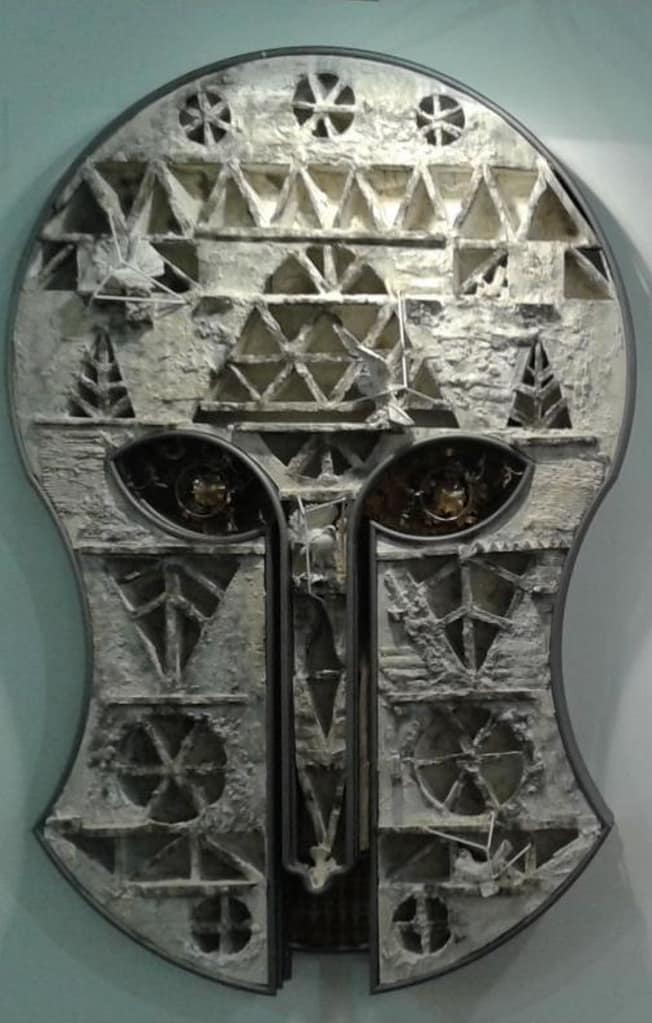 Eine metallische, abstrakte Maskenskulptur mit geometrischen Mustern und Ausschnitten, die zwei markante Augenformen und eine verwitterte, strukturierte Oberfläche in Grau- und Weißtönen aufweist.