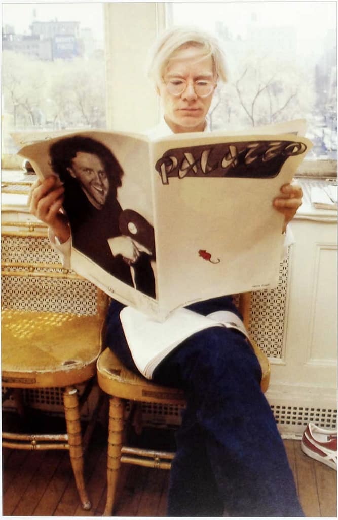 Eine Person mit hellem Haar und Brille sitzt auf einem goldenen Stuhl am Fenster und liest eine große Zeitschrift mit dem Titel „PALAZZO“, auf deren Cover ein Schwarzweißfoto eines lächelnden Mannes ist.