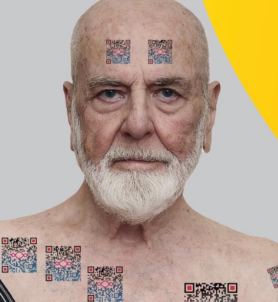 Ein älterer Mann mit weißem Bart und ernstem Gesichtsausdruck steht mit freiem Oberkörper da und trägt mehrere QR-Code-Tattoos auf Stirn, Wangen, Brust und Schultern. Der Hintergrund ist hellgrau mit einer gelben, geschwungenen Form.