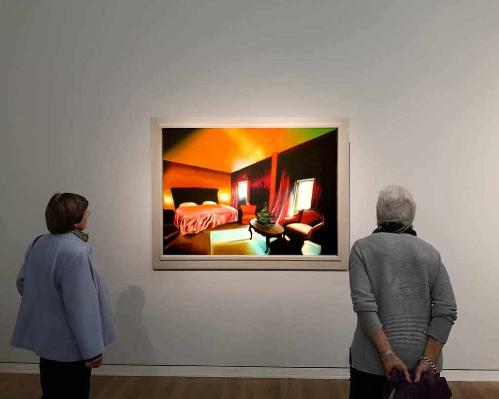 Gemälde Heinz Zolper: Zwei Personen betrachten in einer Galerie ein lebendiges, abstraktes Gemälde. Das Kunstwerk zeigt eine Innenraumszene in kräftigen Farben wie Orange und Rot mit einem Bett, Stühlen und einer Topfpflanze. Die Betrachter scheinen sich mit der Kunst zu beschäftigen.